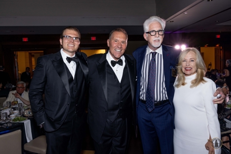 A group of four smiling people, three men and one woman, dressed in formal attire, stand together at an event.